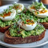 Sliced soft-boiled eggs and fresh microgreens garnish avocado on warm, crisp whole grain toast.